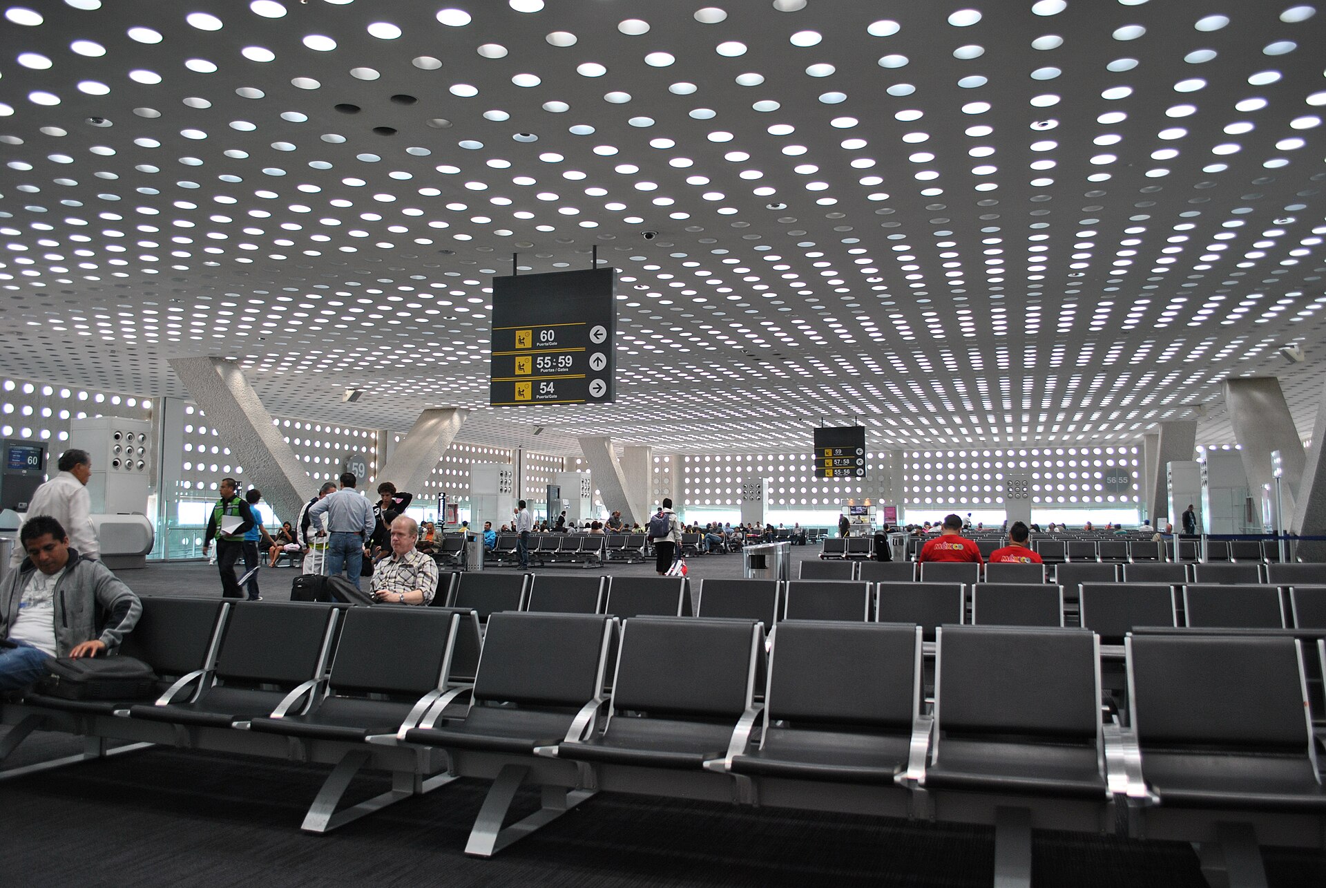 The departures waiting area in Terminal 2 of Mexico City International Airport, with a perforated ceiling of circular lights and bilingual gate signage