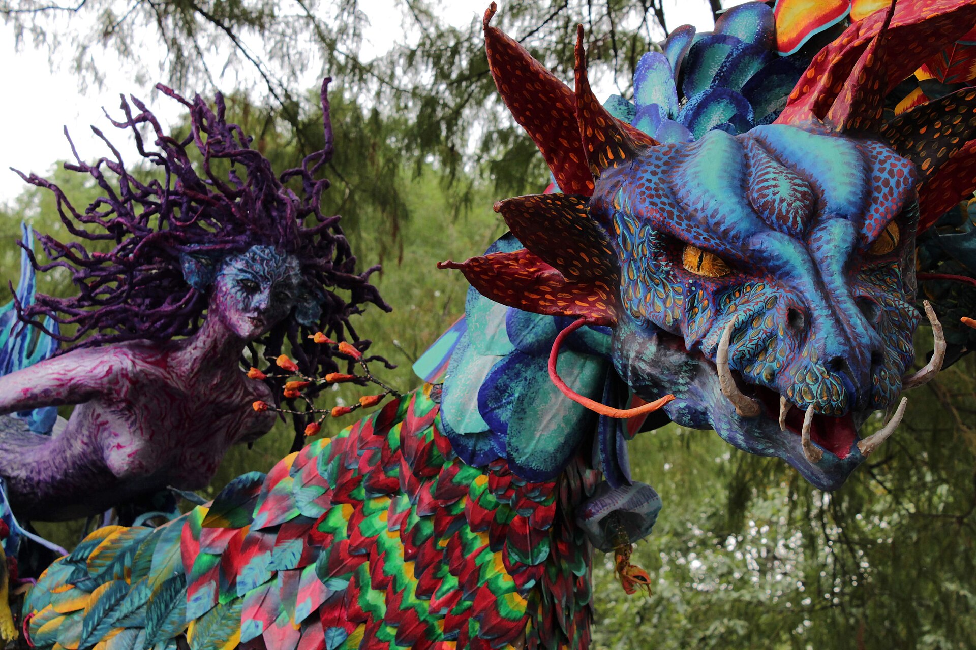 A monumental alebrije sculpture of a blue horned creature with rainbow feathered scales displayed among trees on Paseo de la Reforma in Mexico City