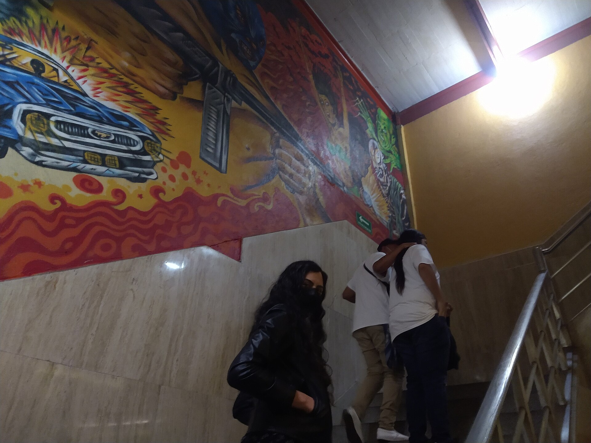 Spectators climbing a staircase inside Arena México past a painted mural of masked luchadores, a car and flames