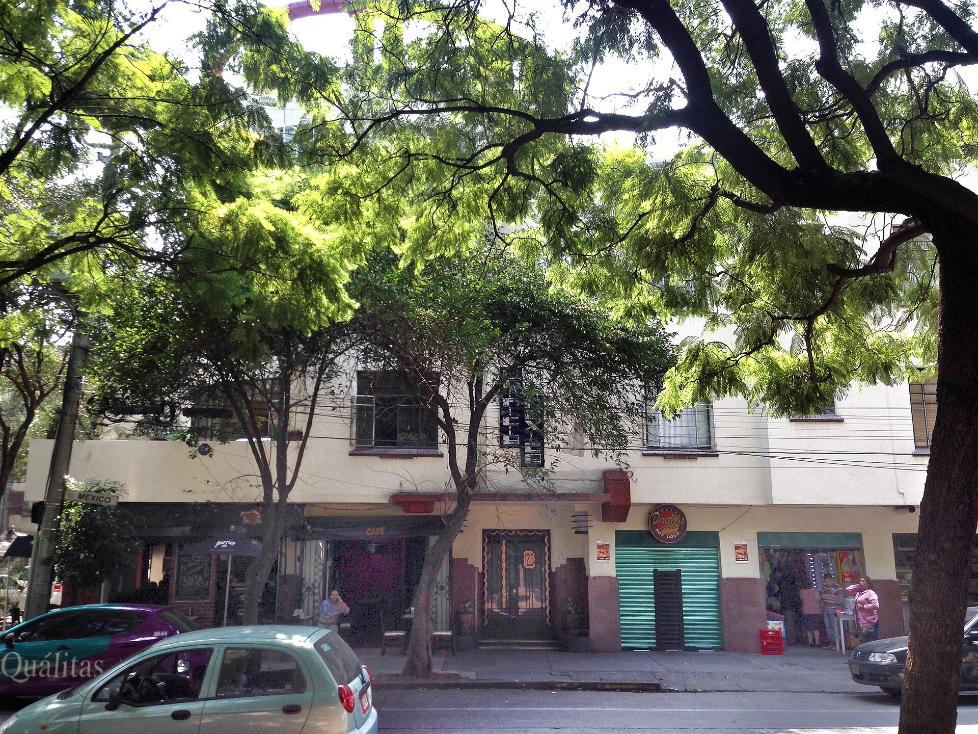 Low-rise cream-coloured buildings with ground-floor cafes and shops on a corner of Avenida Mexico in the Hipodromo section of Condesa, shaded by a large tree