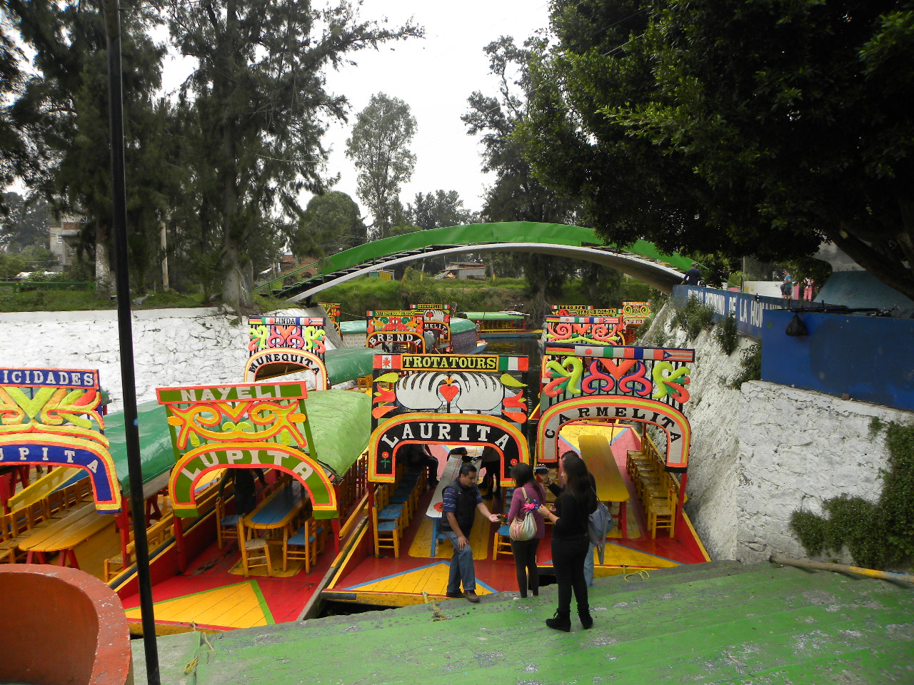 Visitors boarding brightly painted trajinera boats with named arches and wooden chairs at the Las Flores Nativitas landing in Xochimilco