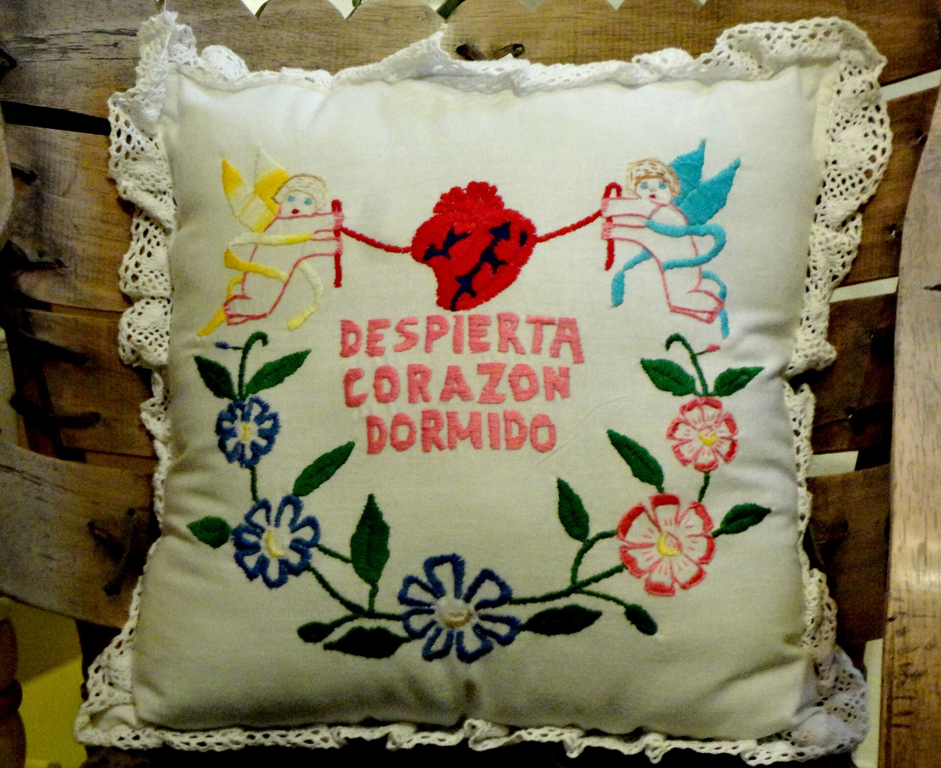 An embroidered cushion made by Frida Kahlo showing a pierced red heart held by two cherubs above the words Despierta corazón dormido, surrounded by embroidered flowers