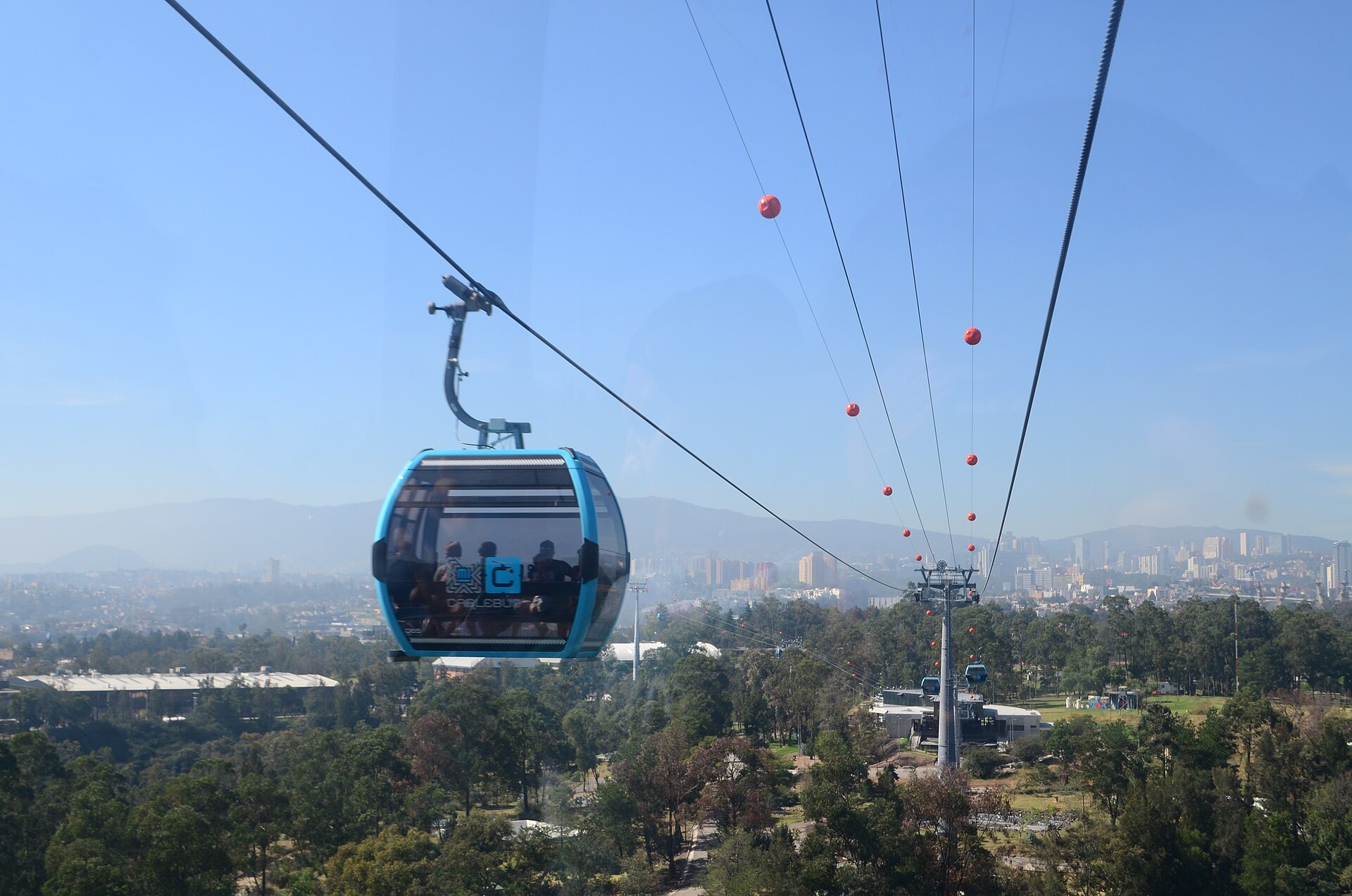 A blue Cablebus gondola carrying passengers above trees and the Mexico City skyline, with the cable line running away towards a station