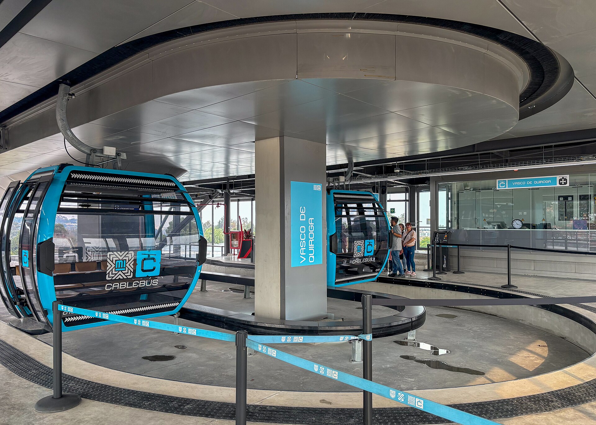 Two blue Cablebús gondolas in the Vasco de Quiroga station in Mexico City, with passengers waiting on the platform