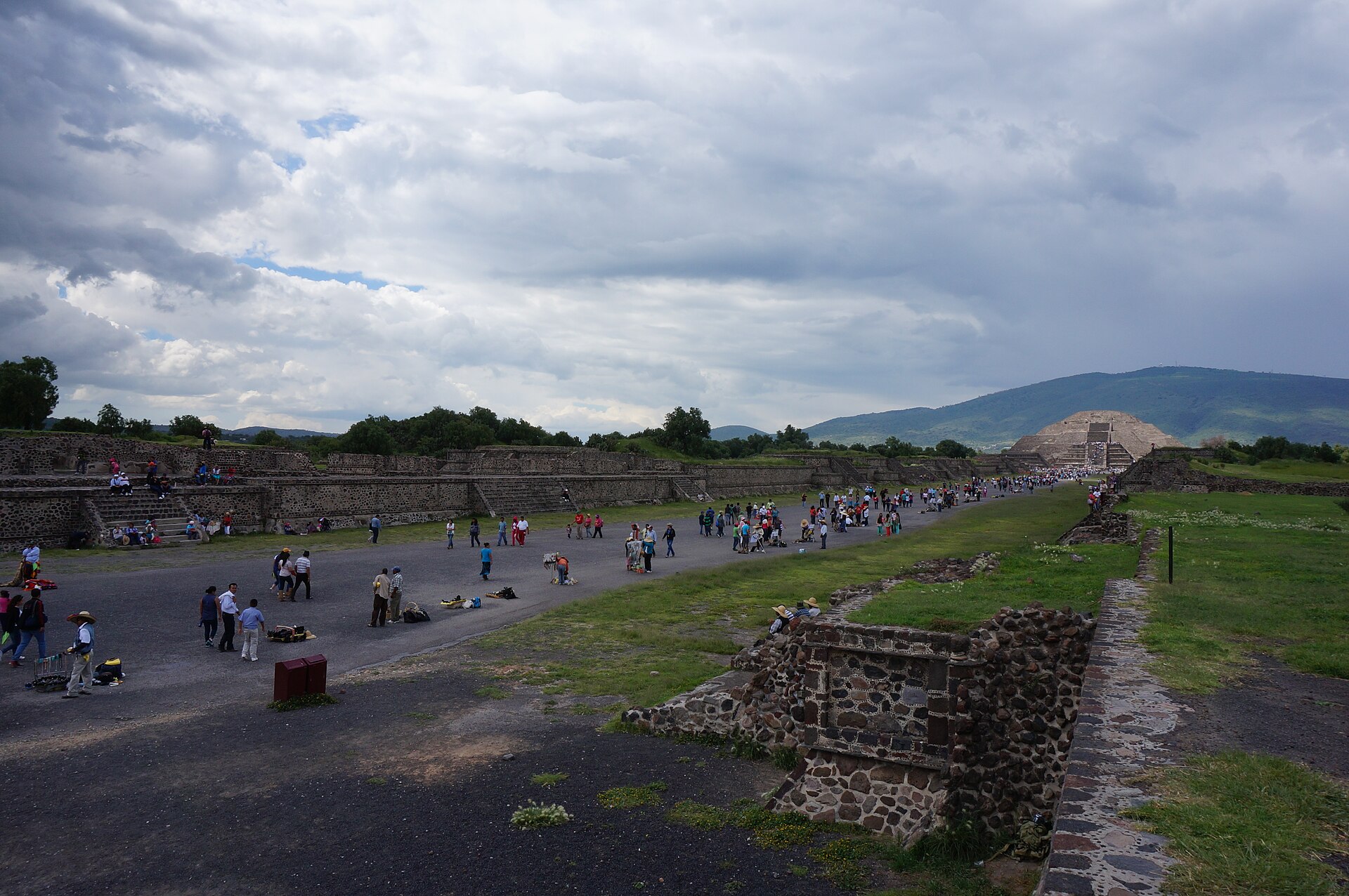 The Avenue of the Dead at Teotihuacan running north between low stone platforms towards the Pyramid of the Moon, with visitors walking under a cloudy sky