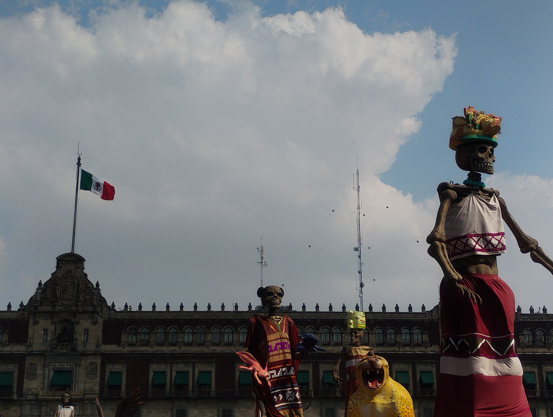 Giant skeleton figures in traditional dress standing in the Zocalo in front of the Palacio Nacional during Dia de Muertos, with the Mexican flag flying behind