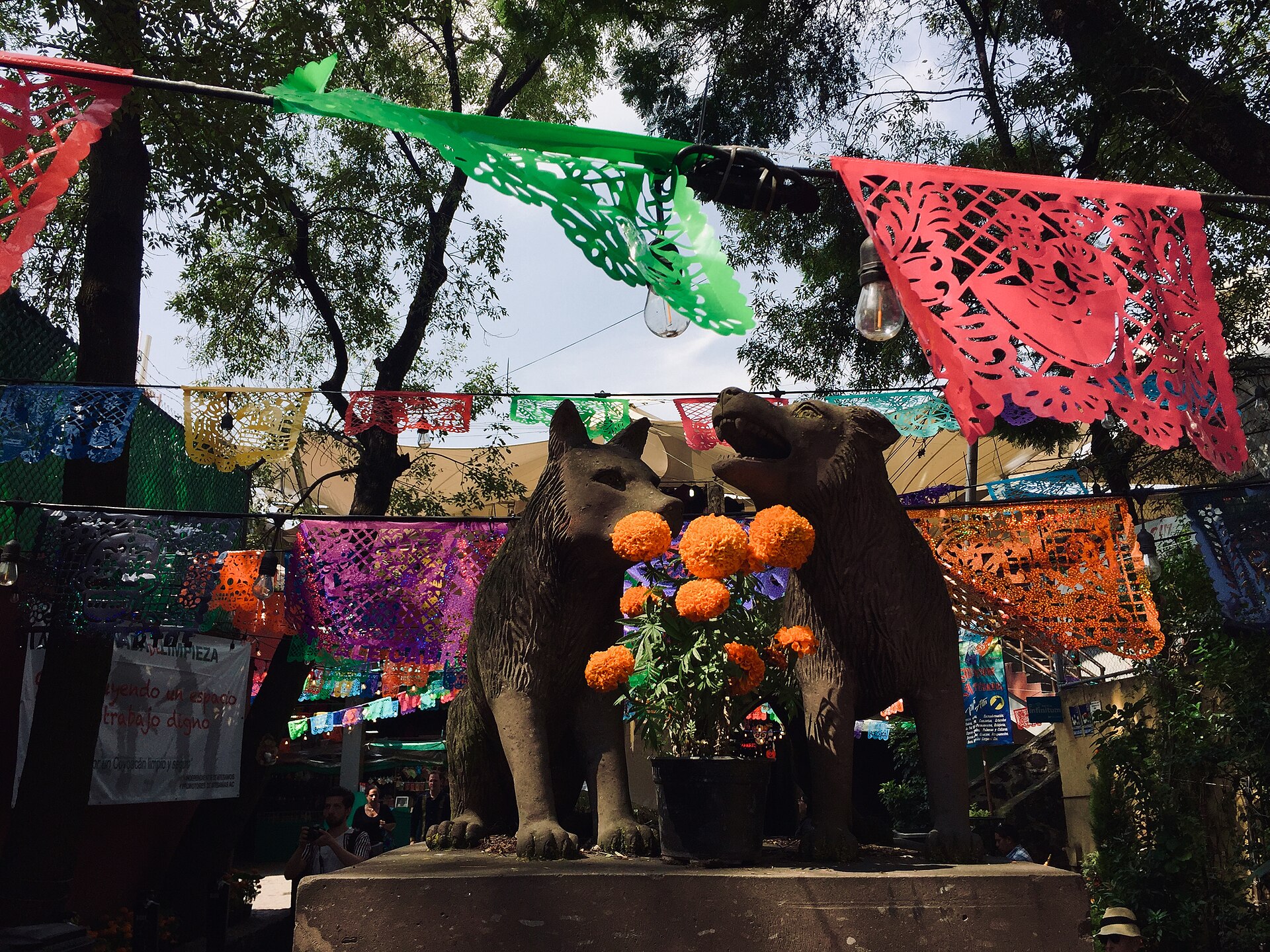 Two stone coyote sculptures on a plinth in Coyoacan, surrounded by marigolds and strings of coloured papel picado