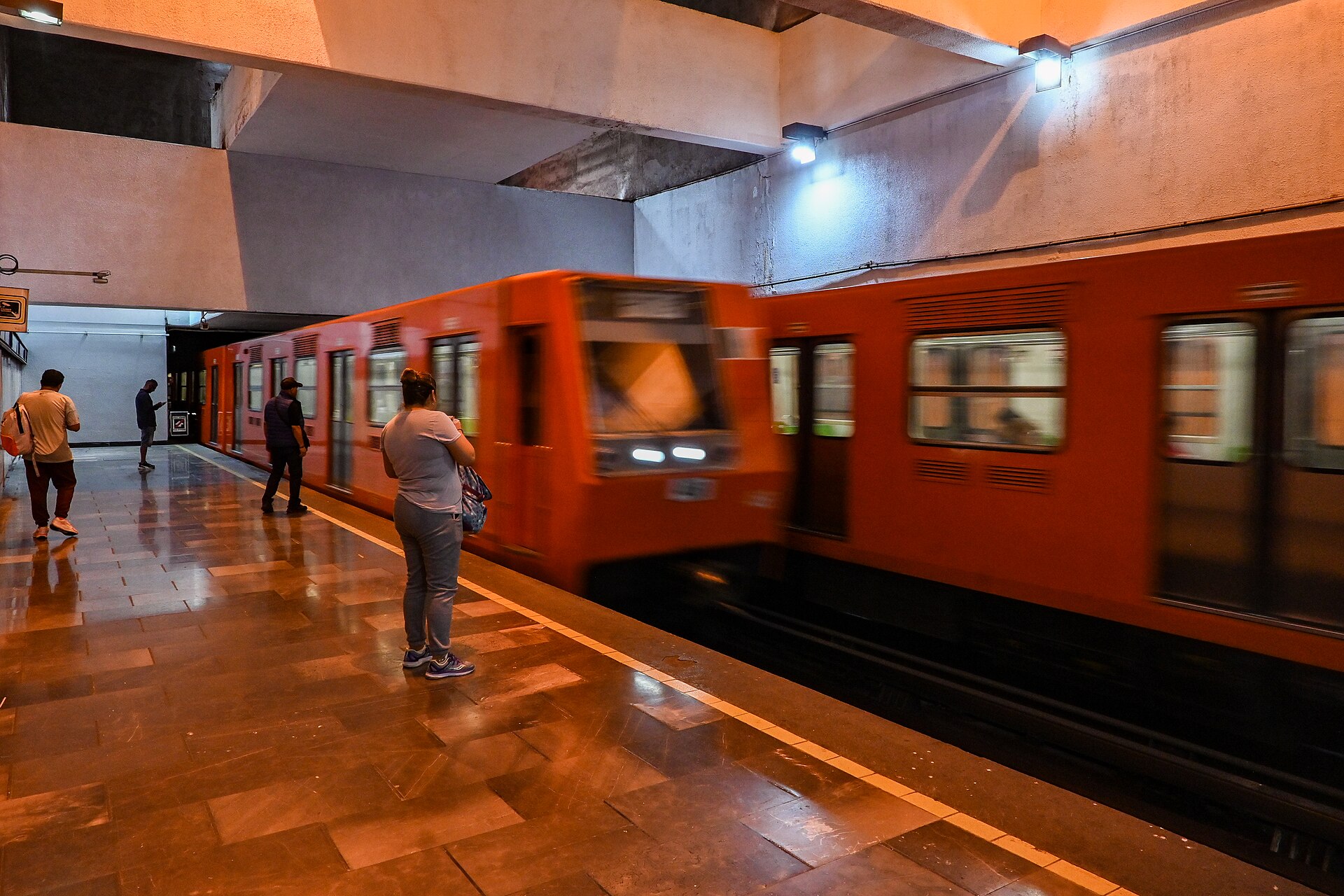 An orange Mexico City Metro train arriving at an underground platform while passengers wait on the polished stone floor