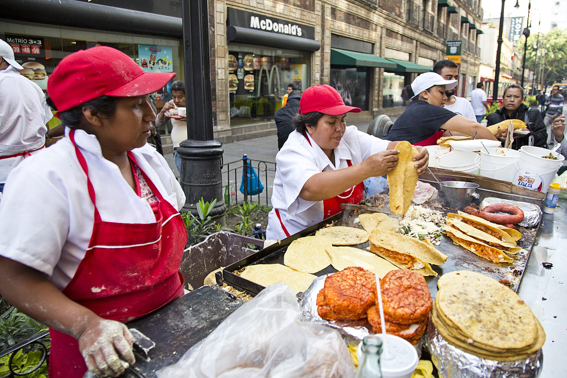 Two vendors in red caps and aprons working a hot griddle piled with quesadillas and tacos at a busy street stand in central Mexico City