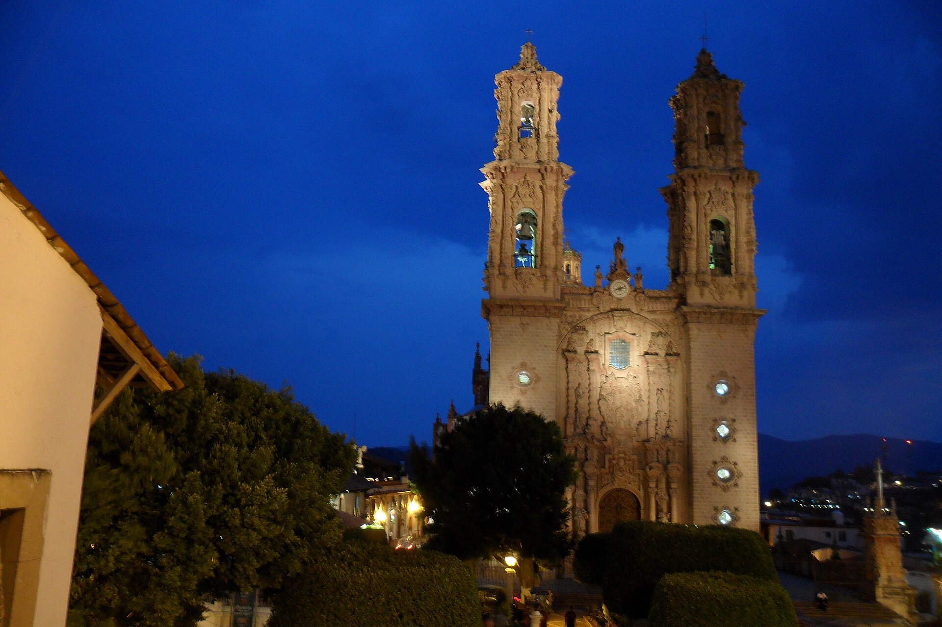 The floodlit churrigueresque twin towers of the Parroquia de Santa Prisca in Taxco at dusk, with the hillside town behind