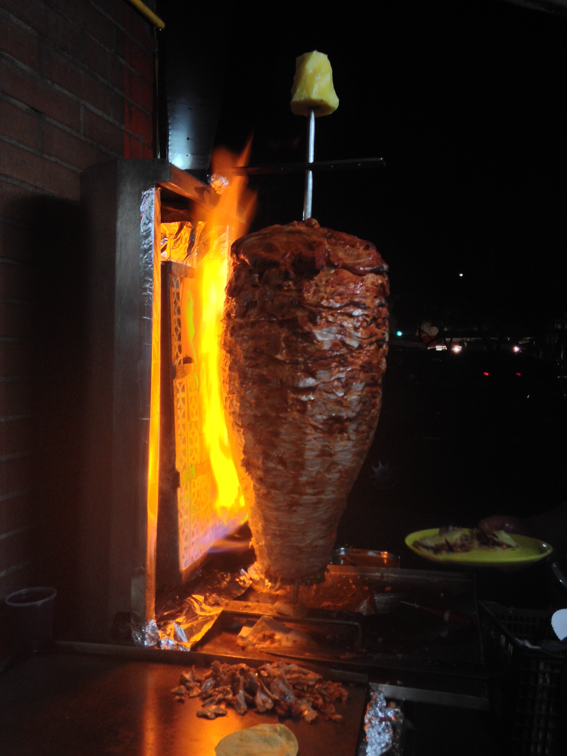 A vertical spit of marinated pork for tacos al pastor turning beside an open gas flame at night, with a whole pineapple fixed at the top