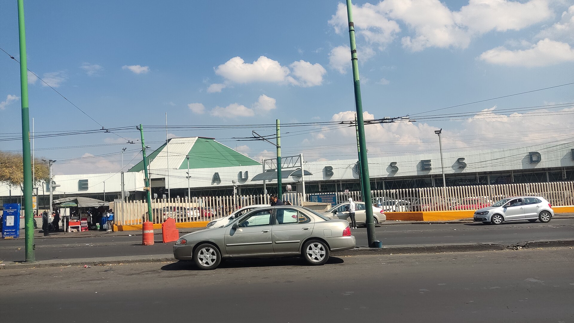 The long, low facade of the Central de Autobuses del Norte in Mexico City with large metal letters spelling Autobuses del Norte, a green pyramid-shaped roof and cars on the road in front