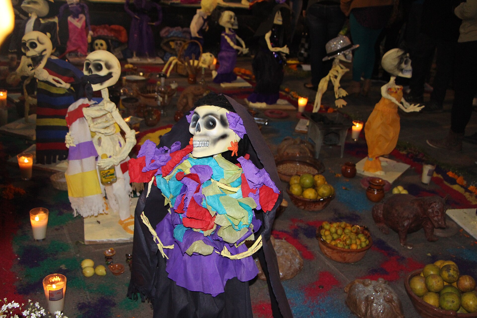 A Day of the Dead ofrenda at night in Coyoacán, with papier-mâché skeleton figures in bright crêpe-paper clothes, lit candles, bowls of fruit and pan de muerto on a coloured sawdust carpet