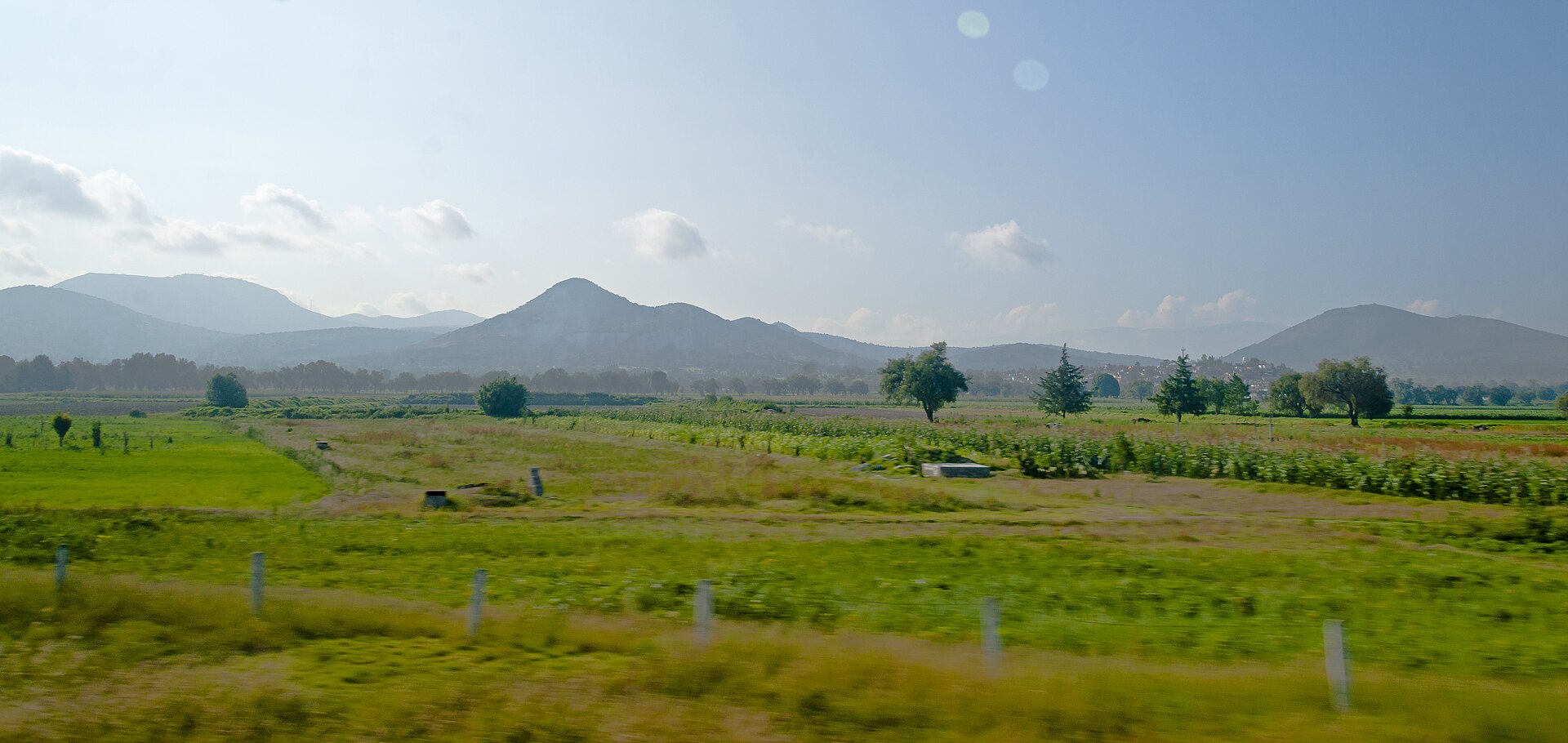 Green farmland and fields of maize with low wooded hills behind, seen from the window of a bus on the highway north-east of Mexico City
