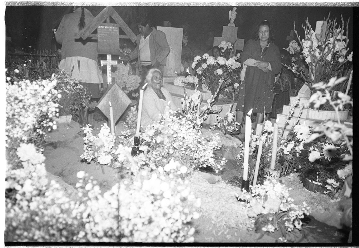 Black and white photograph of the cemetery at Mixquic at night on 2 November 1986, with an elderly woman seated among flowers and candles at a grave and other family members standing nearby