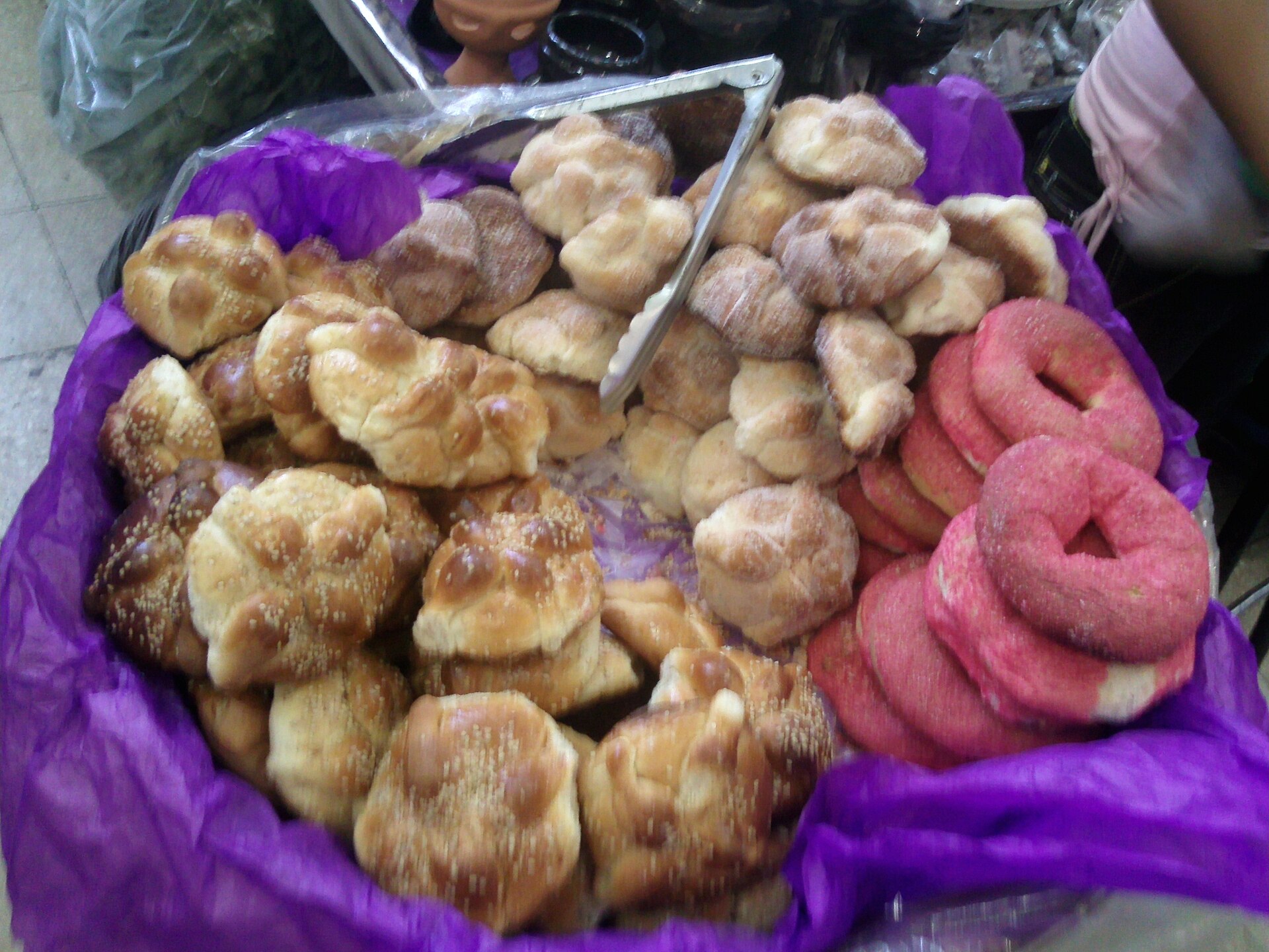 A basket lined with purple tissue paper full of pan de muerto, some glazed and sprinkled with sesame, some dusted with sugar, and pink sugared rings, for sale at a market in Coyoacán