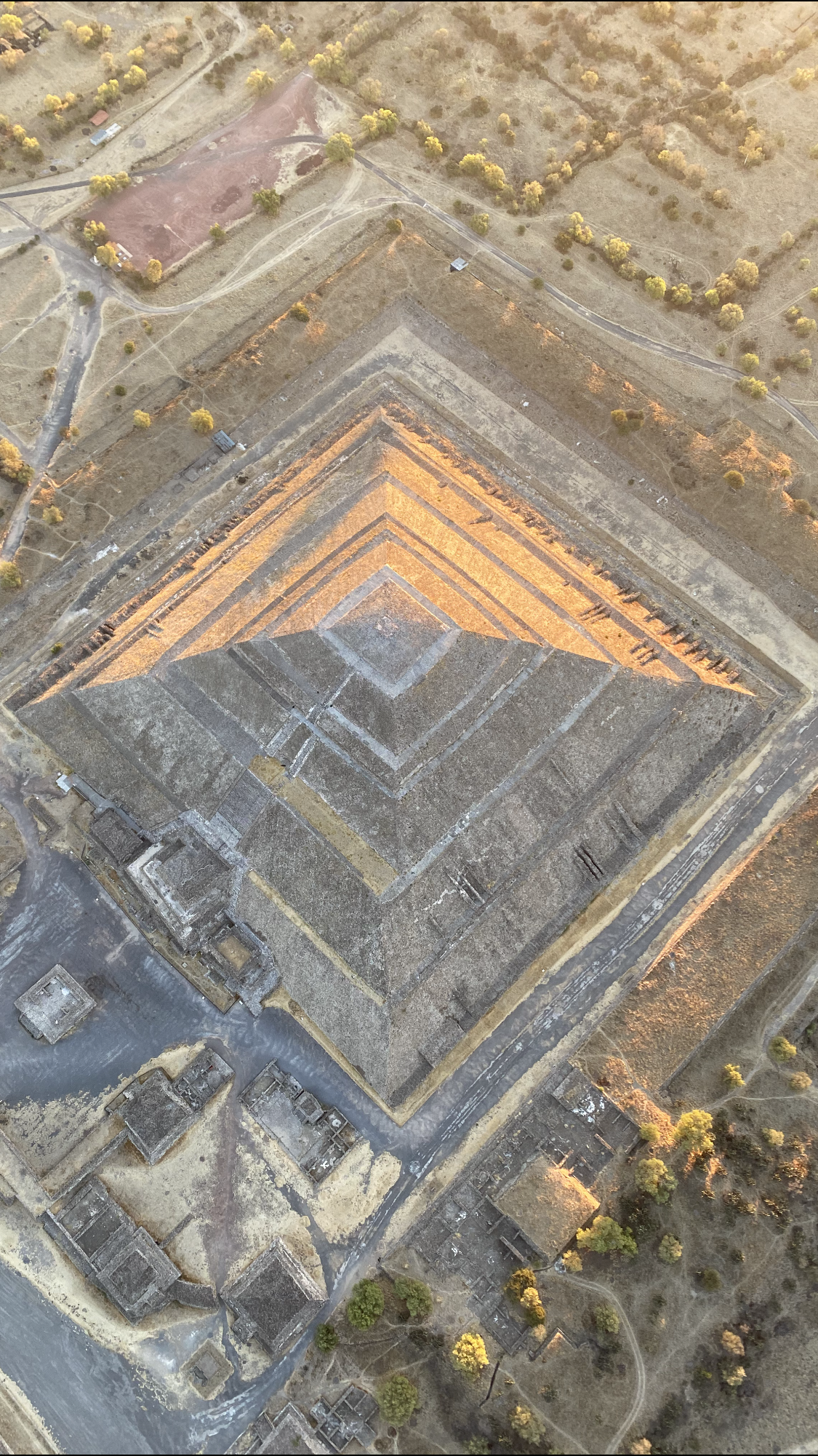 The Pyramid of the Sun seen from directly above at first light, its stepped terraces lit orange on one side and in shadow on the other, with ruined platforms and dirt paths around its base