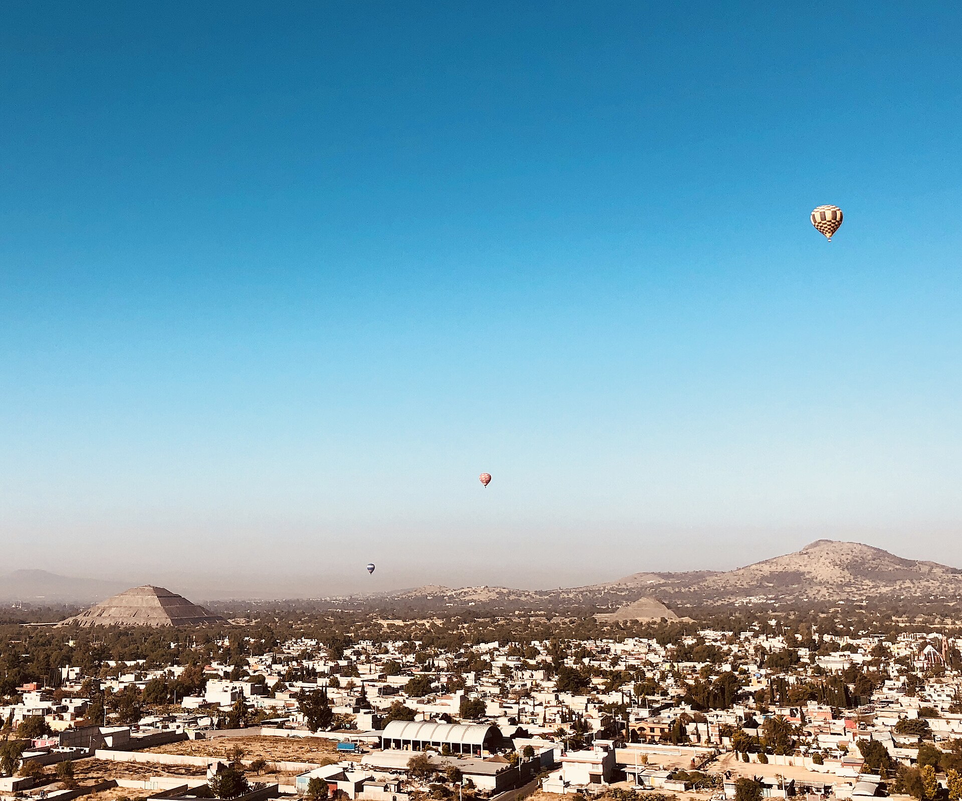 View from a hot air balloon across the flat rooftops of a town towards the Pyramid of the Sun and the smaller Pyramid of the Moon on the horizon, with three more balloons in a clear blue morning sky