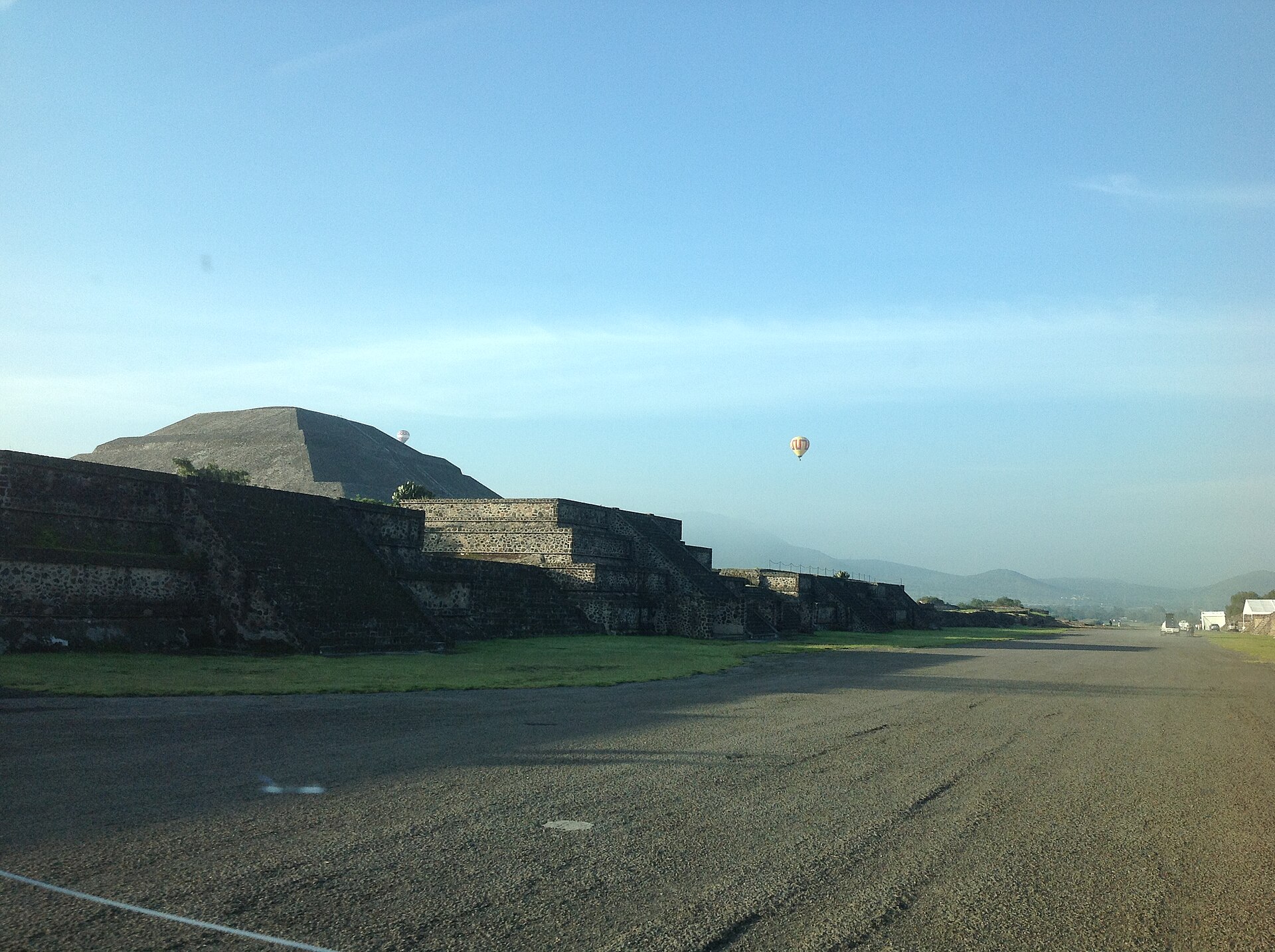 Two hot air balloons in the early morning sky near the Pyramid of the Sun at Teotihuacan, seen past a row of low stone platforms beside a wide gravel road